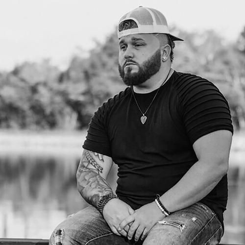 Man with a beard and tattoos sitting by a lake, wearing a cap backwards and a necklace.