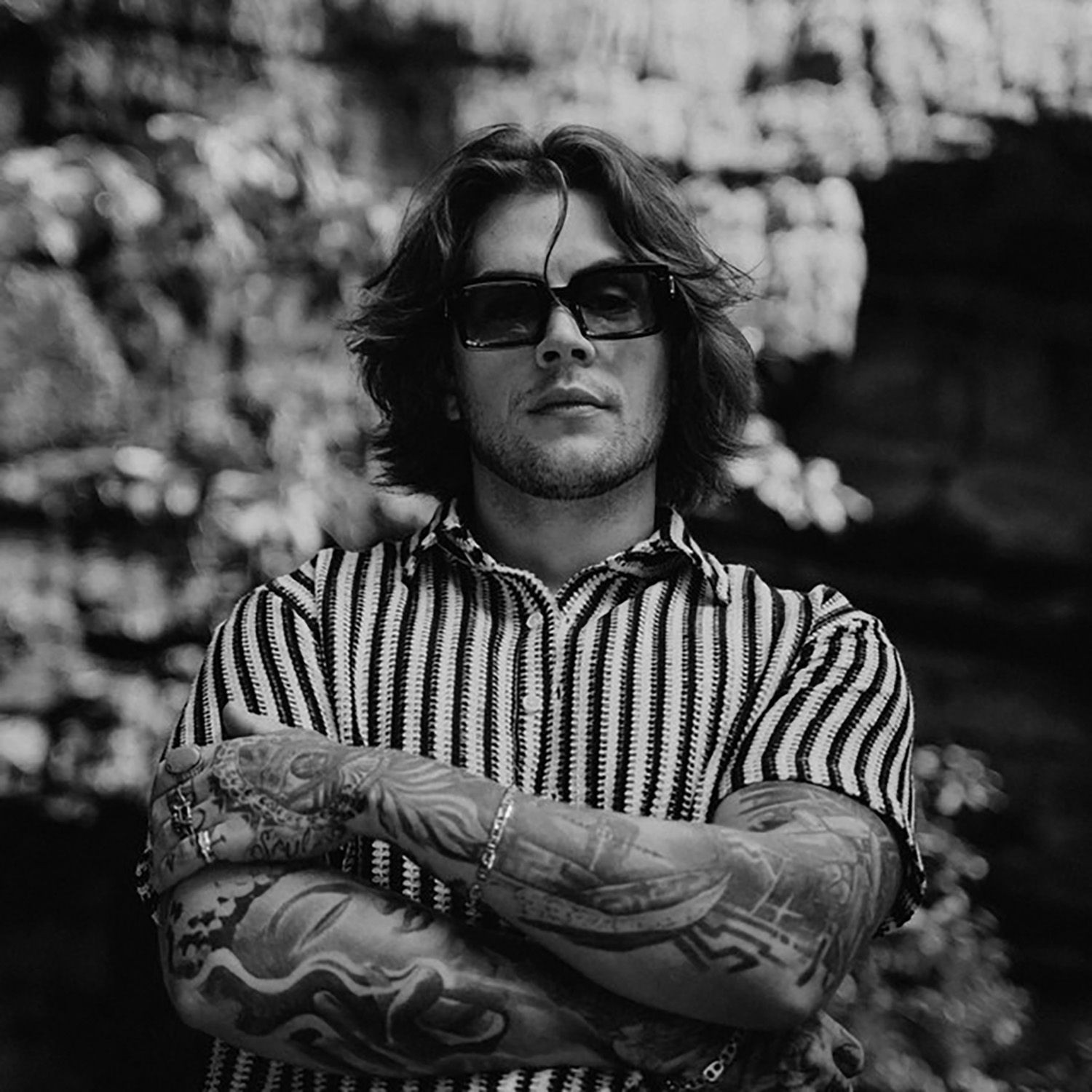 Person with sunglasses and tattoos crossing arms, wearing a striped shirt, in black and white.