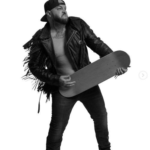 Man in a leather jacket poses with a skateboard like a guitar.