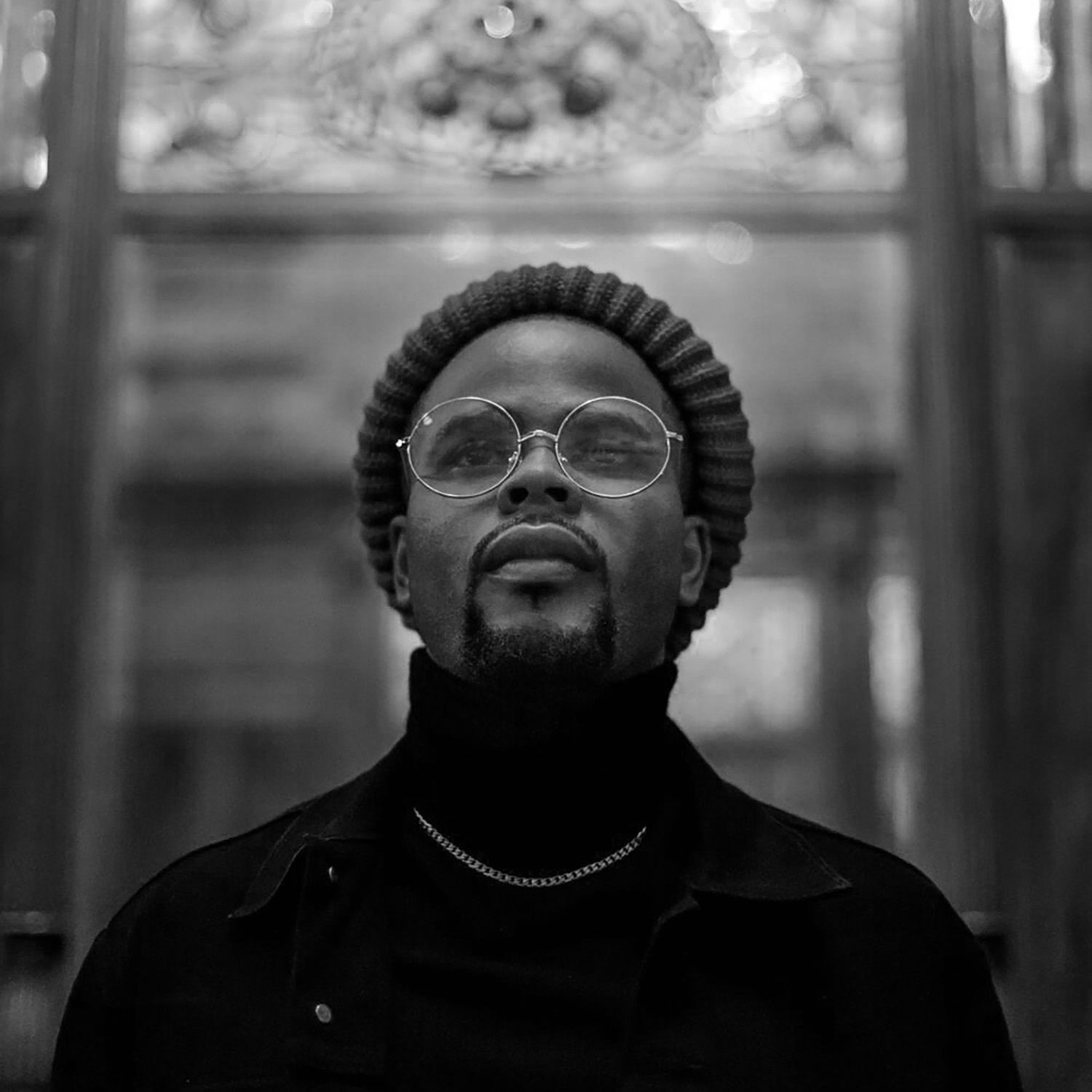 Black and white portrait of a person wearing glasses and a beanie, looking upward.