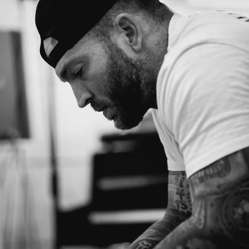 Black and white profile of a man with tattoos, wearing a cap and leaning forward in thought.