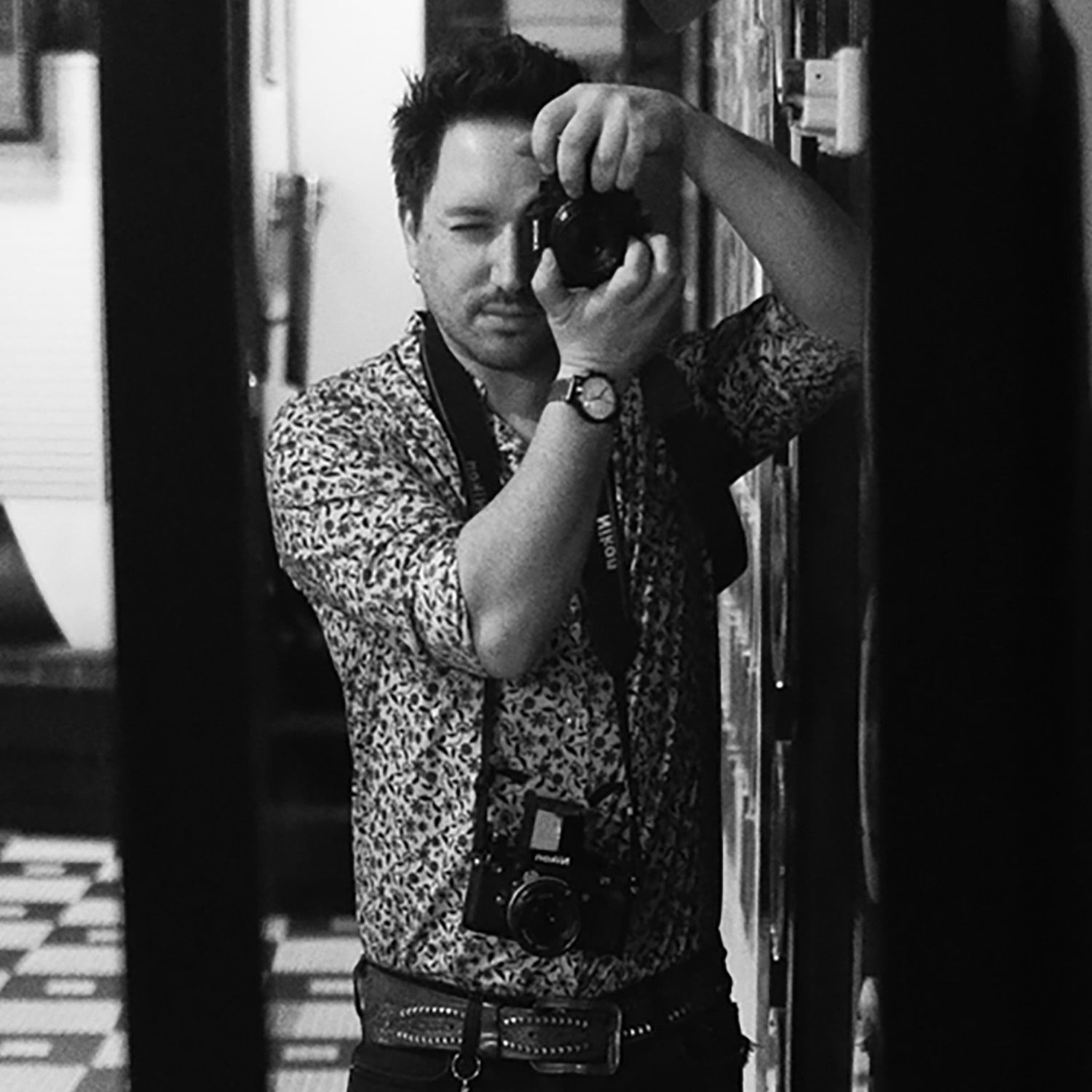 Black and white photo of a man taking a picture with a camera, wearing a patterned shirt.