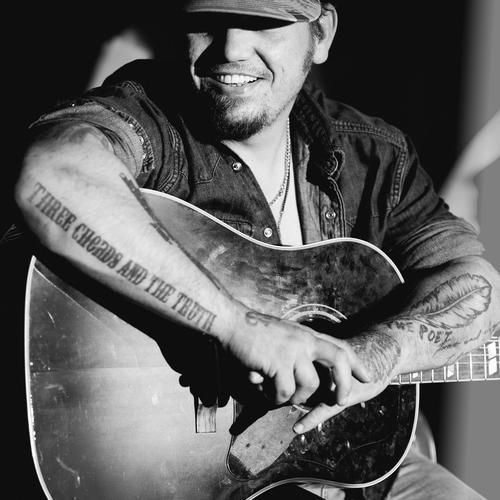 Black and white portrait of a man with tattoos holding an acoustic guitar and smiling.